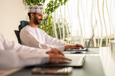 إتمام المهام في المكتب، تطور الشركات العمانية ,الاستعانة بالأجهزة التقنية الحديثة،  العمل الجماعي و التعاون، موظفان عربيان خليجيان عمانيان يرتديان الدشداشة والكمة عمانيان ويقومان باستخدام اللابتوب و التابلت, الجلوس على كرسي مريح