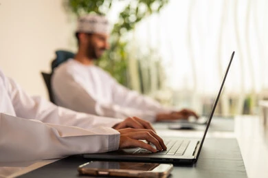 تطور الشركات العمانية , الاستعانة بالأجهزة التقنية الحديثة، إتمام المهام في المكتب، العمل الجماعي و التعاون، موظفان عربيان خليجيان عمانيان يرتديان الدشداشة والكمة عمانيان ويقومان باستخدام اللابتوب و التابلت, الجلوس على كرسي مريح