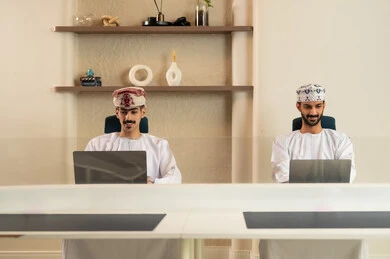 The use of modern technological devices, completing tasks in the office, the development of Omani companies, teamwork and collaboration, two Arab Gulf Omani employees wearing the dishdasha and kumma, using a laptop and a tablet, sitting on a comfortable chair.