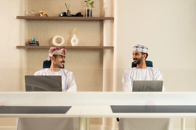 The use of modern technological devices, completing tasks in the office, the development of Omani companies, teamwork and collaboration, two Arab Gulf Omani employees wearing the dishdasha and kumma, using a laptop and a tablet, sitting on a comfortable chair.