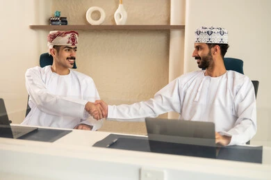 The development of Omani companies, the use of modern technological devices, completing tasks in the office, teamwork and collaboration, two Arab Gulf Omani employees wearing the traditional dishdasha and kumma are using a laptop and a tablet, sitting on a comfortable chair.