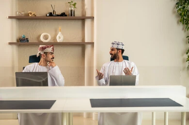 إتمام المهام في المكتب، تطور الشركات العمانية ,الاستعانة بالأجهزة التقنية الحديثة،  العمل الجماعي و التعاون، موظفان عربيان خليجيان عمانيان يرتديان الدشداشة والكمة عمانيان ويقومان باستخدام اللابتوب و التابلت, الجلوس على كرسي مريح