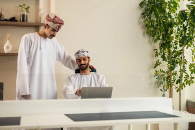 تبادل الخبرات و المعلومات لتحقيق الاهداف،التحفيز المعنوي بين الزملاء,  استخدام الحاسوب المحمول ،تطوير و دراسة المشاريع الجديدة في الشركات العمانية، ايماءات السعادة و السرور, مفهوم التعاون و العمل المشترك, رجلان عربيان خليجيان عمانيان مبتسمان يرتديان الدشداشة و الكمة و يربت أحدهما على كتف الاخر