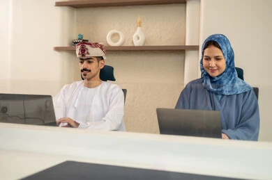 A sophisticated office environment, task division among employees, monitoring daily tasks, completing office work, the concept of project management, developing plans and achieving company goals, an Arab Gulf Omani team sitting at a desk using a laptop.