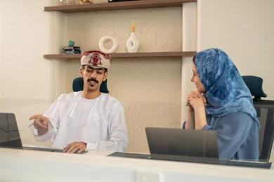 شرح واضح للتاسكات , نقاش و حوار في بيئة مكتبية, تقسيم المهام بين الموظفين, متابعة المهام اليومية، إنجاز الأعمال المكتبية، مفهوم ادارة المشاريع، تطوير الخطط وتحقيق أهداف الشركة، فريق عمل عربي خليجي عماني يجلسون على مكتب يستخدمون الحاسوب المحمول