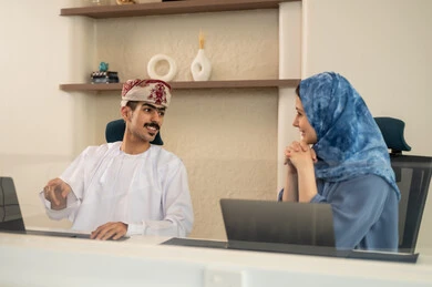 A clear explanation of tasks, discussion and dialogue in an office environment, division of tasks among employees, follow-up on daily tasks, completion of office work, the concept of project management, developing plans and achieving the company's goals, an Arab Gulf Omani team sitting at a desk using a laptop.