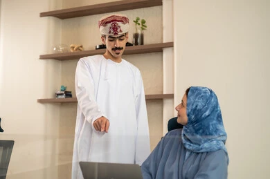 A sophisticated office environment, task division among employees, monitoring daily tasks, completing office work, the concept of project management, developing plans and achieving company goals, an Arab Gulf Omani team sitting at a desk using a laptop.