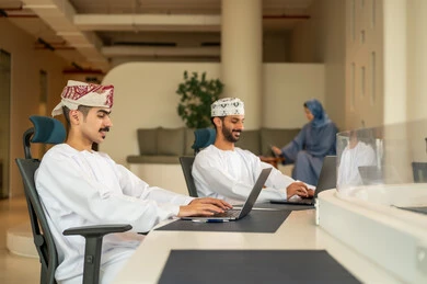 Dividing tasks among employees, following up on daily tasks, completing office work, discussions and debates about projects, developing plans and achieving the company's goals, a Gulf Arab Omani team consisting of two men wearing the dishdasha and kumma using a laptop, and a veiled woman wearing a blue abaya sitting at the desk and using a tablet.
