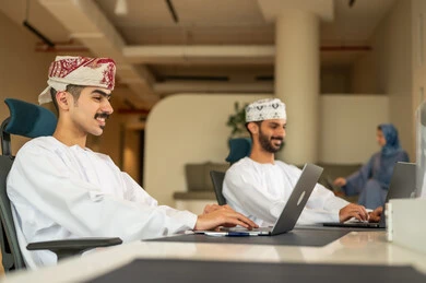 Completing office tasks, dialogues and discussions about projects, dividing tasks among employees, following up on daily tasks, developing plans and achieving the company's goals, a Gulf Arab Omani team consisting of two men wearing dishdasha and kumma using a laptop, and a veiled woman wearing a blue abaya sitting at the desk and using a tablet.