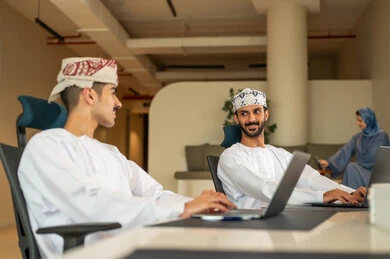 تقسيم المهام بين الموظفين, متابعة المهام اليومية، إنجاز الأعمال المكتبية، الحوارات و النقاشات حول المشاريع، تطوير الخطط وتحقيق أهداف الشركة، فريق عمل عربي خليجي عماني مكون من رجلين يرتديان الدشداشة و الكمة يستخدمون الحاسوب المحمول وسيدة محجبة ترتدي عباءة زرقاء تجلس على المكتب و تستخدم التابلت