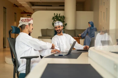 متابعة المهام اليومية، إنجاز الأعمال المكتبية، الحوارات و النقاشات حول المشاريع، تطوير الخطط وتحقيق أهداف الشركة، فريق عمل عربي خليجي عماني مكون من رجلين يرتديان الدشداشة و الكمة يستخدمون الحاسوب المحمول وسيدة محجبة ترتدي عباءة زرقاء تجلس على المكتب و تستخدم التابلت