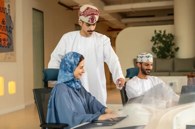 Dividing tasks among employees, following up on daily tasks, completing office work, discussions and debates about projects, developing plans and achieving the company's goals, a Gulf Arab Omani team consisting of two men wearing the dishdasha and kumma using a laptop, and a veiled woman wearing a blue abaya sitting at the desk and using a tablet.