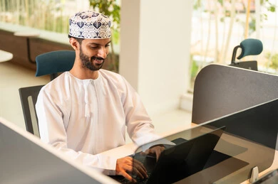 Managing and organizing business affairs via a laptop, pressing the keyboard keys, integrating work with technology and tech, an Arab Gulf Omani man wearing a dishdasha and kumma using a laptop, the concept of completing tasks remotely, a comfortable office work environment.