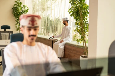 التعاون و متابعة الاعمال, نمط الحياة في الشركات العمانية ، دمج التقنيات المتطورة فى العمل ، تدريب الموظفين على سياسات العمل لتحقيق الأهداف في المكتب داخل مقر الشركة، فريق عربي خليجي عماني مكون من رجلين يرتديان الدشداشة والكمة , انجاز المهام من خلال اللابتوب و التابلت