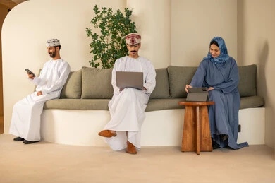 Companies and offices in the Sultanate of Oman, managing and organizing business affairs, teamwork and collaboration, a Gulf Arab Omani team consisting of two men wearing the dishdasha and kumma and a veiled woman wearing a blue abaya, using a laptop, tablet, and mobile phone, gestures of integration and focus.