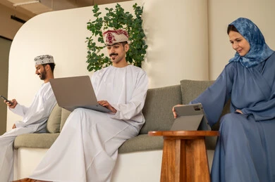 إدارة شؤون الأعمال و تنظيمها، العمل الجماعي و التعاون، الشركات و المكاتب في سلطنة عمان، فريق عربي خليجي عماني مكون من رجلان يرتديان الدشداشة والكمة وسيدة محجبة ترتدي العباءة الزرقاء و يقومون باستخدام اللابتوب و التابلت والجوال, ايماءات الاندماج و التركيز