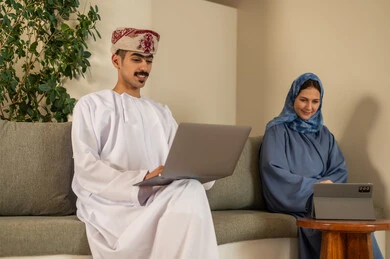 الشركات و المكاتب في سلطنة عمان، إدارة شؤون الأعمال و تنظيمها، العمل الجماعي و التعاون، فريق عربي خليجي عماني مكون من رجل يرتدي الدشداشة والكمة  وسيدة محجبة ترتدي العباءة الزرقاء و يقومون باستخدام اللابتوب و التابلت, ايماءات الاندماج و التركيز