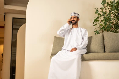 إجراء مكالمة صوتية عبر الهاتف المحمول، القيام بمحادثة عمل من خلال الهاتف, تصفح وسائل التواصل الاجتماعي، إتمام المهام عبر الإنترنت ، إتمام مهام العمل في المكتب، موظف عربي خليجي عماني يرتدي الدشداشة والكمة ويقوم باستخدام الجوال