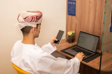 Looking at expressions of integration and interaction, performing daily tasks within the company headquarters, an Arab Gulf Omani employee wearing a dishdasha and kumma is using a laptop and holding a mobile phone in his hand, integrating technology and modern devices with practical life.