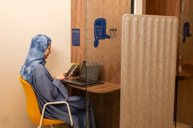 A Saudi Arabian Gulf female employee wearing a blue abaya and hijab is using a laptop and holding a mobile phone in her hand, performing her daily tasks inside the company headquarters, with expressions of happiness and joy, integrating technology and modern devices with professional life.