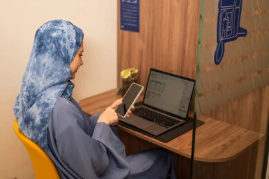 Performing daily tasks inside the company headquarters, with expressions of happiness and joy, an Arab Gulf Saudi female employee wearing a blue abaya and hijab is using a laptop and holding a mobile phone in her hand, integrating technology and modern devices with practical life. Performing daily tasks inside the company headquarters, with expressions of happiness and joy, an Arab Gulf Saudi female employee wearing a blue abaya and hijab is using a laptop and holding a mobile phone in her hand, integrating technology and modern devices with practical life.