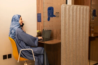 Performing daily tasks inside the company headquarters, with expressions of happiness and joy, an Arab Gulf Saudi female employee wearing a blue abaya and hijab is using a laptop and holding a mobile phone in her hand, integrating technology and modern devices with practical life.