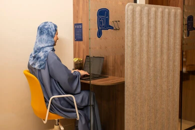 Performing the daily task inside the company headquarters, with expressions of happiness and joy, an Arab Gulf Saudi female employee wearing a blue abaya and hijab is using a laptop, integrating technology and modern devices with practical life.