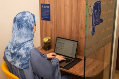 Performing the daily task inside the company headquarters, with expressions of happiness and joy, an Arab Gulf Saudi female employee wearing a blue abaya and hijab is using a laptop, integrating technology and modern devices with practical life.
