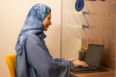 أداء المهمة اليومية داخل مقر الشركة, النظر بتعبيرات السعادة و السرور, موظفه عربيه خليجيه سعودية ترتدي العباءة الزرقاء والحجاب تقوم باستخدام الحاسوب المحمول , دمج التقنية و الأجهزة الحديثة مع الحياة العملية