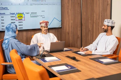 Management and task division, a Gulf Arab Omani team conducting a business meeting inside the conference room at the company's headquarters, explaining and analyzing charts, using a laptop, integrating modern technological devices with office work, studying statistical data sheets, developing projects and investments.