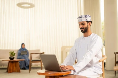 Teamwork and collaboration, companies and offices in the Kingdom of Saudi Arabia, managing and organizing business affairs, an Arab Gulf Omani team consisting of a man wearing a thobe and a headscarf and a woman in a hijab wearing a blue abaya, using a laptop and a tablet, gestures of integration and focus.