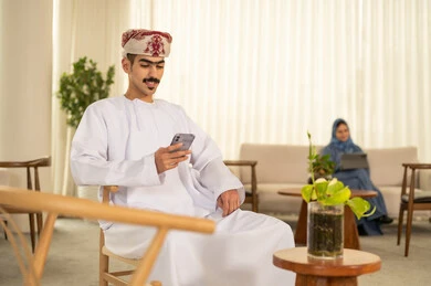 العمل عن بعد، استخدام الأجهزة التكنولوجية الحديثة, رجل عربي خليجي عماني مبتسم يرتدي الدشداشة و الكمة يتصفح مواقع التواصل الاجتماعي عبر الجوال ، امراه سعودية تستخدم الجهاز اللوحي