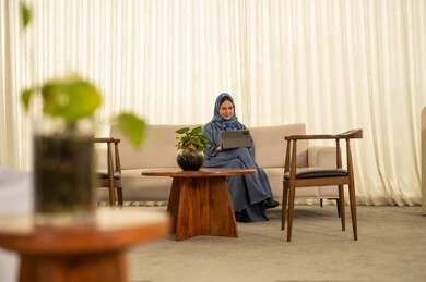 Looking at expressions of happiness and joy, performing daily tasks inside the company headquarters, an Arab Gulf Omani employee wearing a blue abaya and hijab is using a tablet, integrating technology and modern devices with practical life.