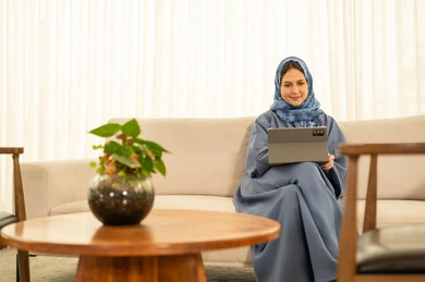 موظفه عربيه خليجيه عمانيه ترتدي العباءة الزرقاء والحجاب تقوم باستخدام التابلت, دمج التقنية و الأجهزة الحديثة مع الحياة العملية، أداء المهمة اليومية داخل مقر الشركة, النظر بتعبيرات السعادة و السرور