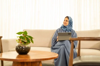 Performing the daily task inside the company headquarters, looking with expressions of happiness and joy, an Arab Gulf Omani female employee wearing a blue abaya and hijab is using a tablet, integrating technology and modern devices with practical life.