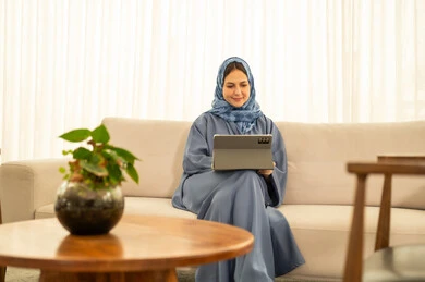 An Arab Gulf Omani female employee wearing a blue abaya and hijab is using a tablet, integrating technology and modern devices with practical life, performing daily tasks inside the company headquarters, with expressions of happiness and joy.