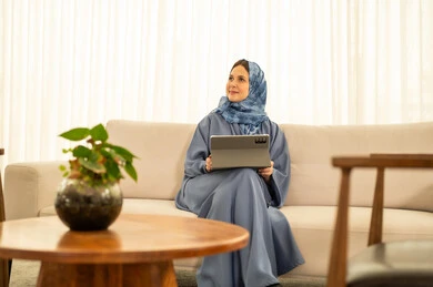 Looking at expressions of happiness and joy, performing daily tasks inside the company headquarters, an Arab Gulf Omani employee wearing a blue abaya and hijab is using a tablet, integrating technology and modern devices with practical life.