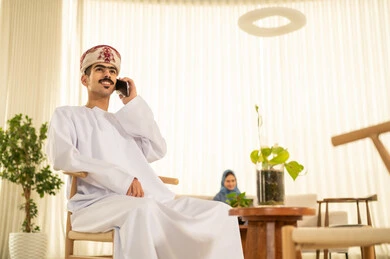 إجراء مكالمة صوتية عبر الهاتف المحمول، القيام بمحادثة عمل من خلال الهاتف, تصفح وسائل التواصل الاجتماعي، إتمام المهام عبر الإنترنت ، إتمام مهام العمل في المكتب، موظف عربي خليجي عماني يرتدي الدشداشة والكمة ويقوم باستخدام الجوال