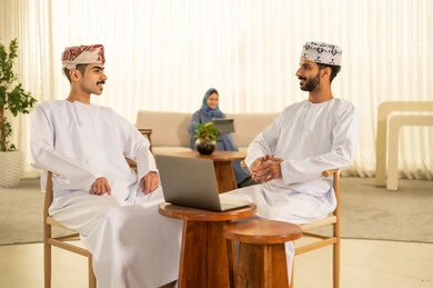 Task division among employees, monitoring daily tasks, integrating advanced technologies into daily life, completing office work, dialogues and discussions about projects, developing plans and achieving the company's goals, a Gulf Arab Omani team consisting of two men wearing the dishdasha and kumma using a tablet, and a veiled woman wearing a blue abaya sitting at the desk and using a tablet.