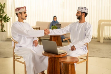 The handshake, greeting, and reception, the concept of cooperation and joint work, expressions and gestures of happiness and joy, two smiling Arab Gulf men from Oman wearing the dishdasha and kumma, one sitting at the desk in front of the laptop and shaking hands with the other. The handshake, greeting, and reception, the concept of cooperation and joint work, expressions and gestures of happiness and joy, two smiling Arab Gulf men from Oman wearing the dishdasha and kumma, one sitting at the desk in front of the laptop and shaking hands with the other.