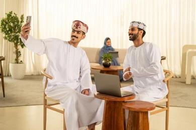 التقاط صور السيلفي التذكارية عبر الجوال, مفهوم التعاون والعمل المشترك, تعابير وايماءات السعادة والسرور، رجلان عربيان خليجيان عمانيان مبتسمان يرتديان الدشداشة و الكمة يجلسان على المكتب أمام اللابتوب