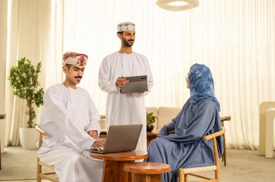 Task division among employees, monitoring daily tasks, integrating advanced technologies into daily life, completing office work, dialogues and discussions about projects, developing plans and achieving the company's goals, a Gulf Arab Omani team consisting of two men wearing the dishdasha and kumma using a tablet, and a veiled woman wearing a blue abaya sitting at the desk and using a tablet.