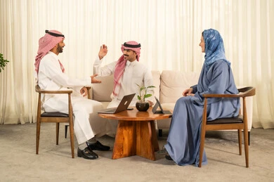 الحوارات و النقاشات حول المشاريع، دمج التقنيات الحديثة فى العمل ،تطوير الخطط وتحقيق أهداف الشركة، التعاون و المشاركة في إنجاز الأعمال، فريق عمل عربي خليجي سعودي مكون من رجلين يرتديان الزي التقليدي والشماغ و سيدة محجبة ترتدي عباءة زرقاء يستخدمون اللابتوب و الجهاز اللوحي, مصافحة و سلام حار بين الزملاء