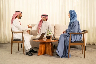 دمج التقنيات الحديثة فى العمل ، الحوارات و النقاشات حول المشاريع، تطوير الخطط وتحقيق أهداف الشركة، التعاون و المشاركة في إنجاز الأعمال، فريق عمل عربي خليجي سعودي مكون من رجلين يرتديان الزي التقليدي والشماغ و سيدة محجبة ترتدي عباءة زرقاء يستخدمون اللابتوب و الجهاز اللوحي, مصافحة و سلام حار بين الزملاء