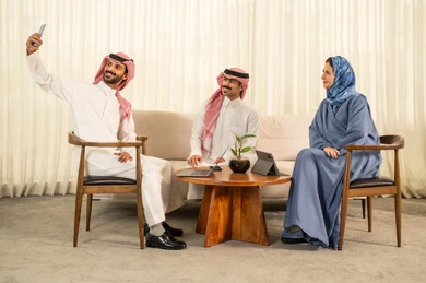 توثيق اللحظات السعيدة ، التقاط صور السيلفي التذكارية عبر الجوال، الحوارات و النقاشات حول المشاريع، تطوير الخطط وتحقيق أهداف الشركة، التعاون و المشاركة في إنجاز الأعمال، فريق عمل عربي خليجي سعودي مكون من رجلين يرتديان الزي التقليدي والشماغ و سيدة محجبة ترتدي عباءة زرقاء تجلس امام الجهاز اللوحي,