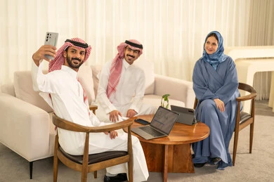 التقاط صور السيلفي التذكارية عبر الجوال، الحوارات و النقاشات حول المشاريع، تطوير الخطط وتحقيق أهداف الشركة، التعاون و المشاركة في إنجاز الأعمال، فريق عمل عربي خليجي سعودي مكون من رجلين يرتديان الزي التقليدي والشماغ و سيدة محجبة ترتدي عباءة زرقاء تجلس امام الجهاز اللوحي