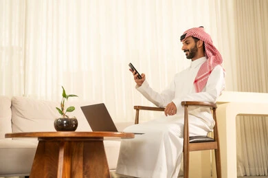 Using the laptop to send emails, preparing and reviewing performance reports, conducting business feasibility studies, integrating modern and advanced technologies with work, administrative office professions and jobs, an Arab Gulf Saudi man wearing traditional attire and a ghutrah sits at the desk in front of the laptop holding a mobile phone in his hand.