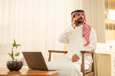 إعداد و مراجعة تقارير الأداء، دراسة جدوى الأعمال، استخدام اللابتوب لإرسال البريد الإلكتروني، دمج التقنيات الحديثة و المتطورة مع العمل, مهن و أعمال مكتبية إدارية, رجل عربي خليجي سعودي يرتدي الزي التقليدي والشماغ يجلس على المكتب امام جهاز اللابتوب ويحمل بيده الهاتف المحمول