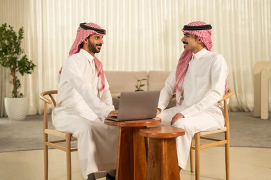 Understanding and cooperation among colleagues to achieve the company's goals, the concept of business management, completing tasks through modern devices, following up on the work strategy, two Arab Gulf men from Saudi Arabia wearing traditional attire and the ghutrah sitting in the office and working on the laptop.