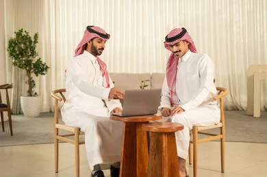 The concept of business management, understanding and cooperation among colleagues to achieve the company's goals, completing tasks through modern devices, following the work strategy, two Arab Gulf men from Saudi Arabia wearing traditional attire and shemagh sitting in the office and working on a laptop.